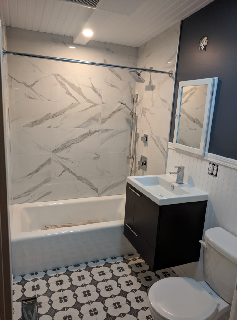 A beautiful bathroom remodel with a modern black vanity, white wooden siding and dark charcoal paint, plus decorative gray and white floor tiles, and marble patterned tiles in the shower.