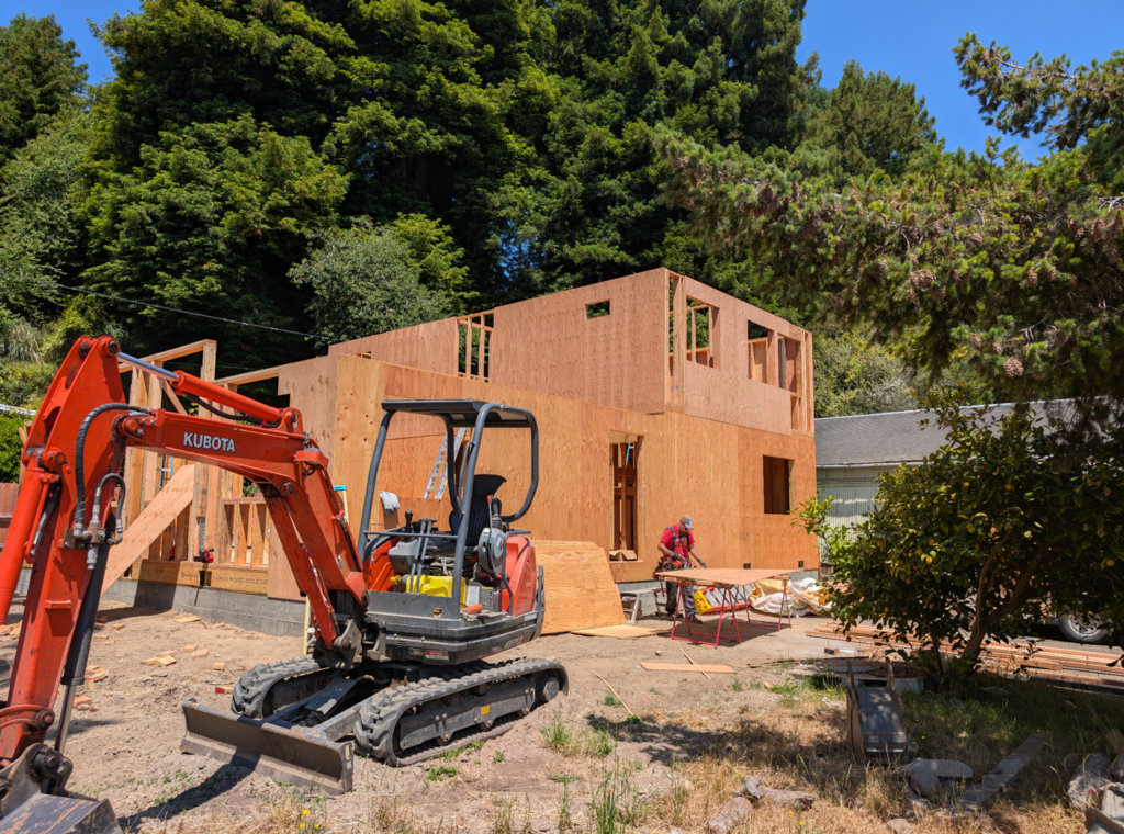 A excavator helps clear the foundation for a new house built by Ultimate Building Solutions. The house is starting to shape with wooden framing as a worker prepares the next pieces.