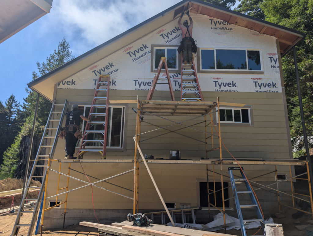A worker stands on a ladder on top of scaffolding to add siding to the second story of a newly built house on a sunny day.