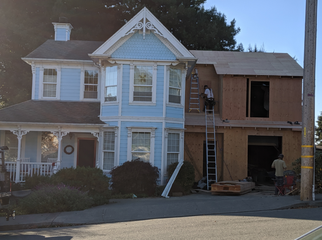 Exterior view from the street of the Victorian remodel. Ladders are leaning against the walls and workers are adding siding to the addition.