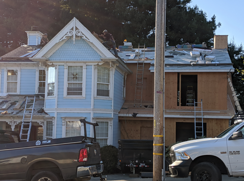 Exterior street view of the Victorian remodel, a large portion of the house is being framed with plywood and the roof is under construction. Workers are removing old shingles from the original roof.