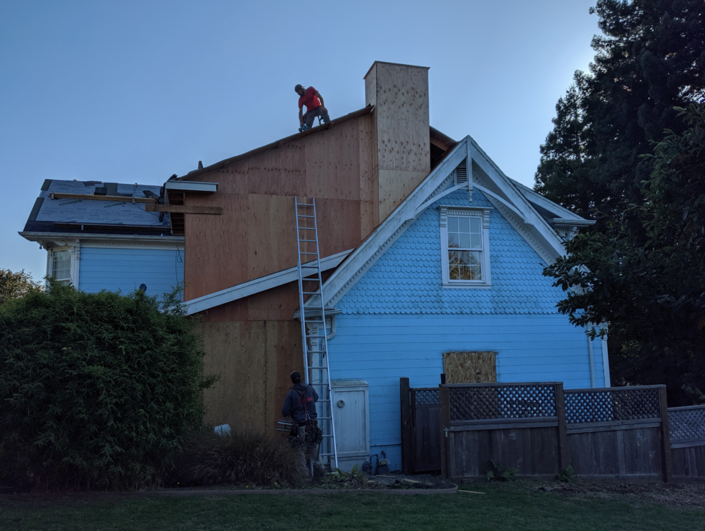 Exterior view of the Blue Victorian remodel with a ladder leaning against the house and a worker fixing the roof.